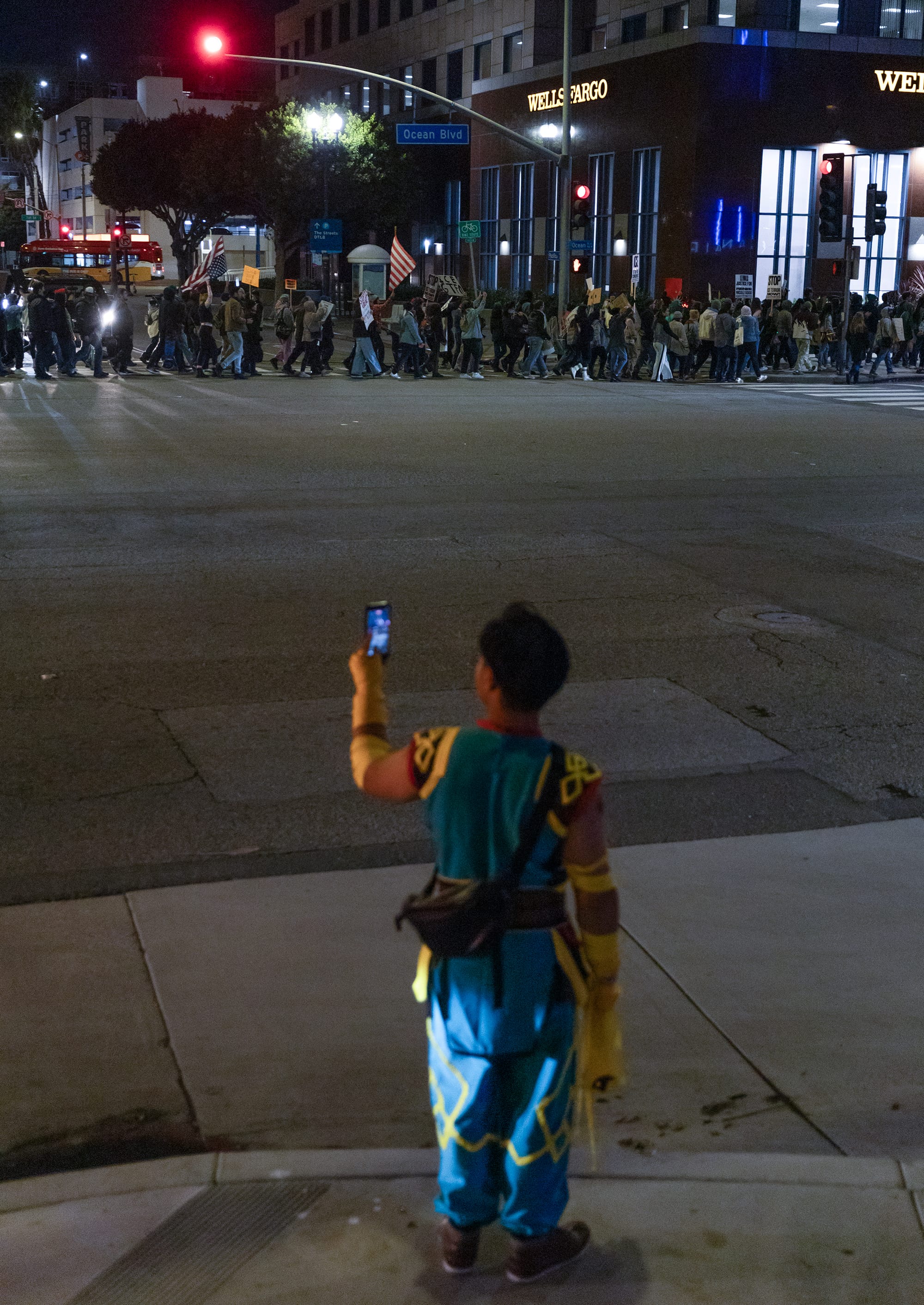 A lone person in a blue costume films a line of people marching across the street.