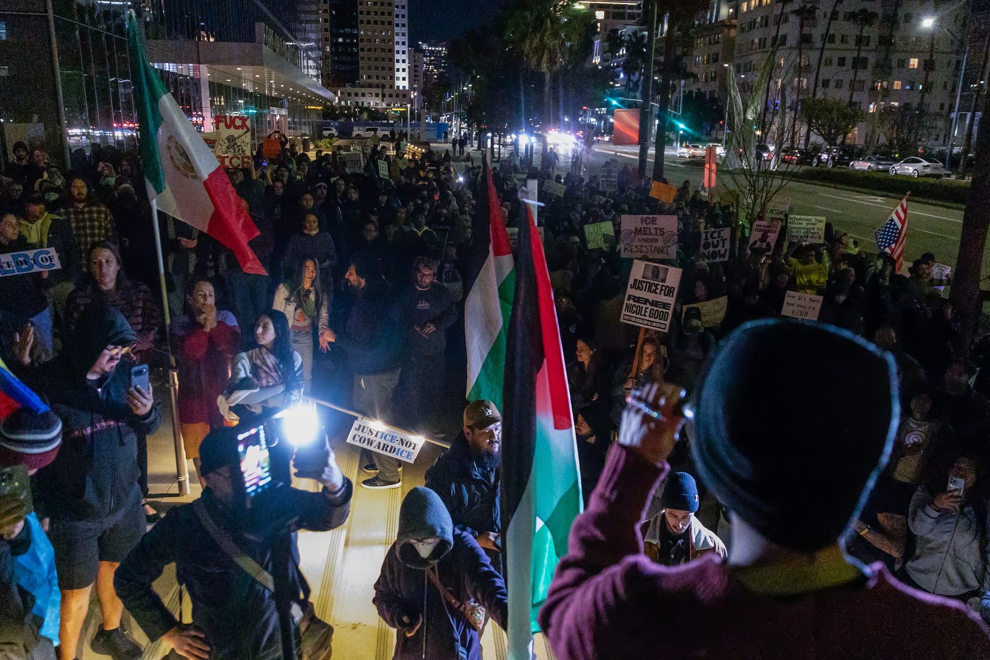 Hundreds of people stand together in a downtown at night.