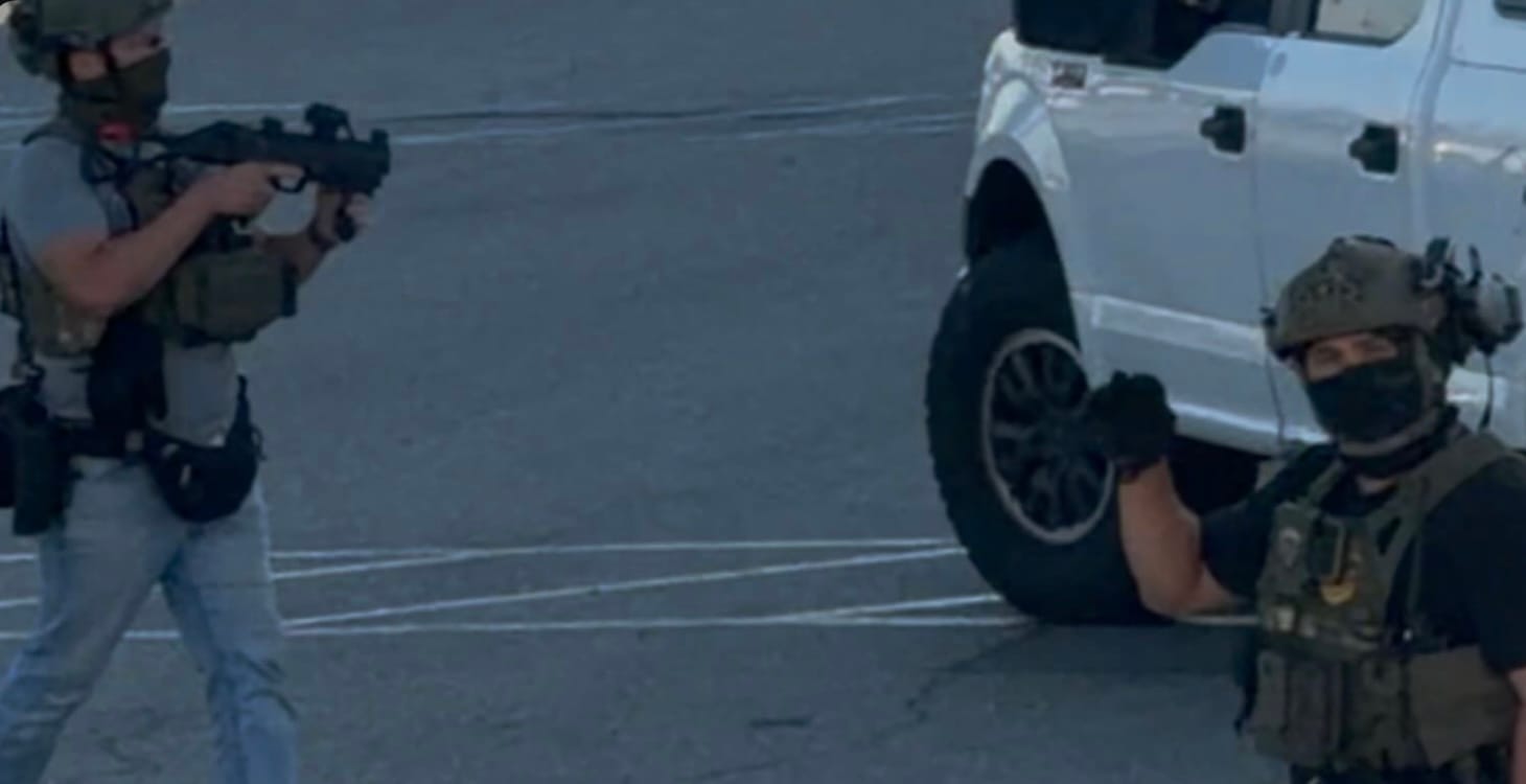 A man wearing body armor and a mask holds a gun near a white truck while another masked man holds up his fist.