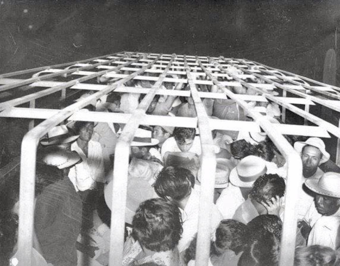 Black and white photo of a few dozen people in a large cage.