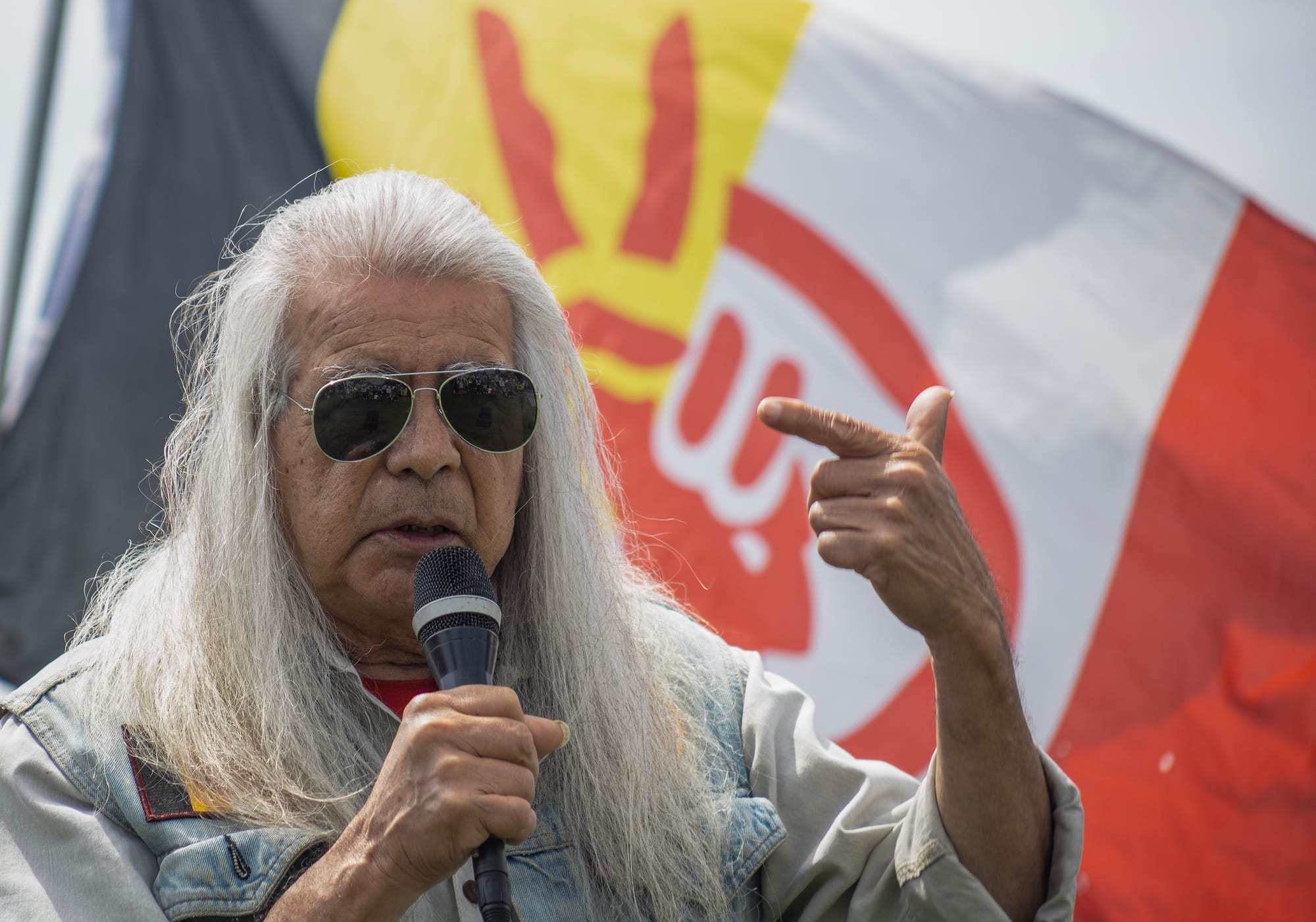 A person with long white hair and sunglasses speaks into a microphone.