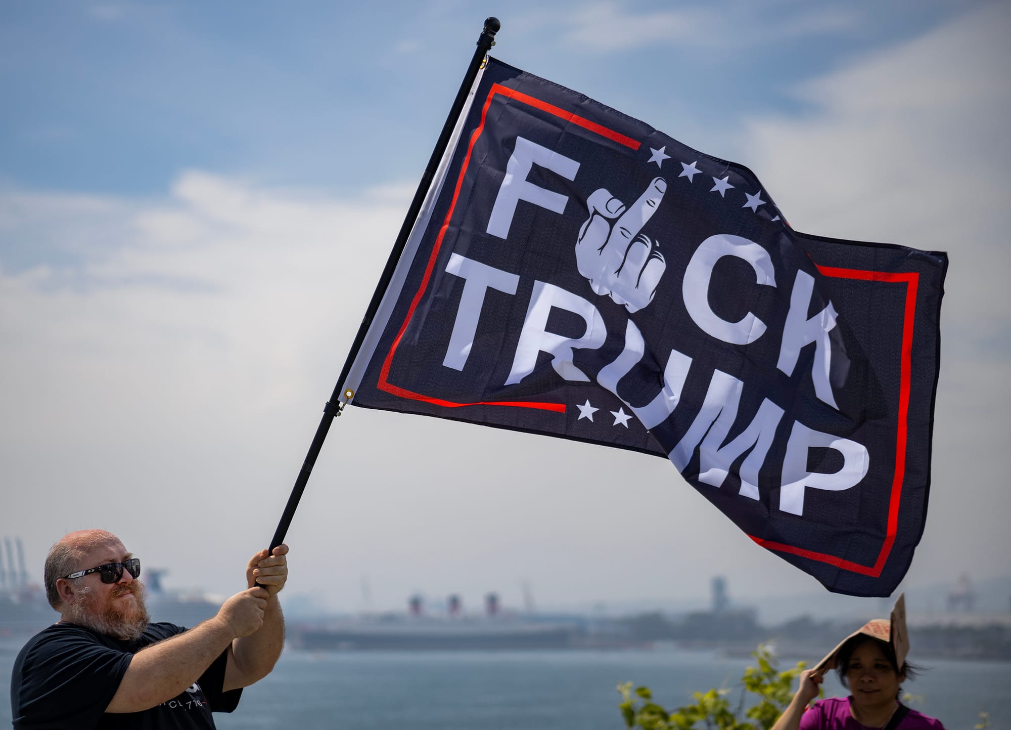 A man waves a flag saying "Fuck Trump" in front of a harbor.