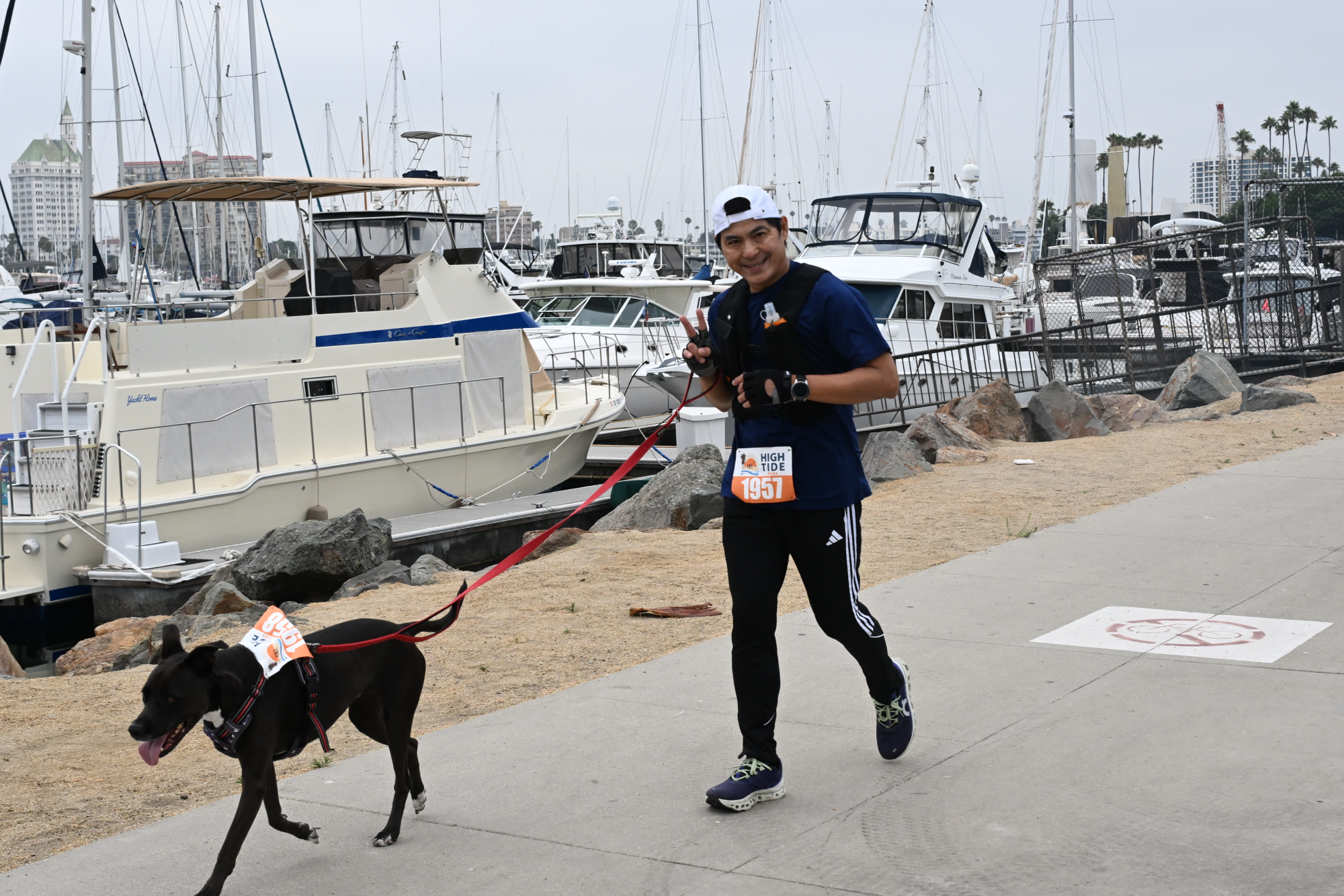 A man is jogging with a black dog on a red leash near a marina.