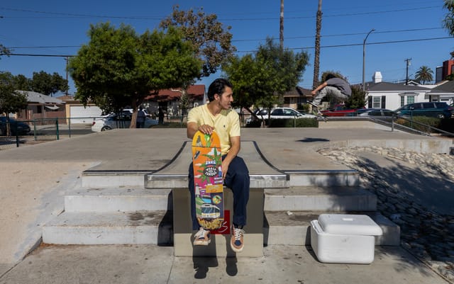 A man wearing a yellow shirt holds a colorful skateboard deck while sitting at a concrete skatepark.