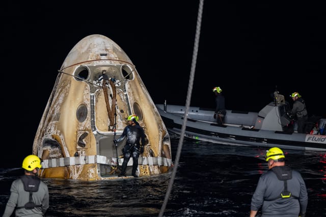 People wearing yellow helmets and gray and black clothing work near a brownish space capsule sitting in water.