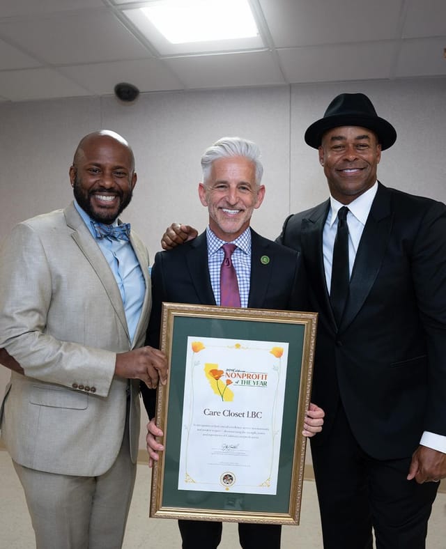 Three men in suits stand while the man in the center holds an award in a gold frame.