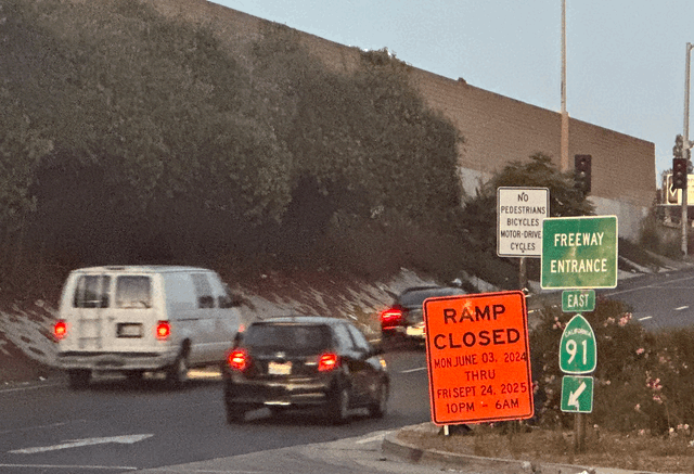 A van and two cars drive on a freeway on-ramp next to orange, green and white signs.