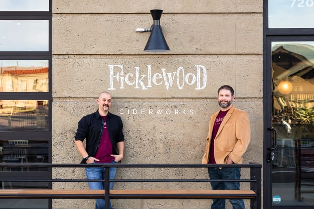 Two men stand in front of a concrete wall with the words "Ficklewood Ciderworks" painted in white on it.