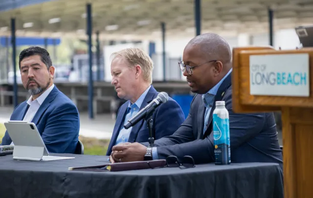 Three men wearing blue suits without ties sit at a blue table next to a lectern labeled "city of Long Beach."