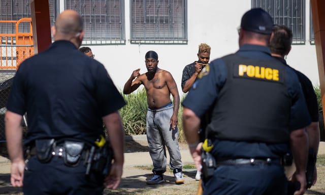 I man wearing gray sweats and no shirt points toward police officers who have their backs to the camera.