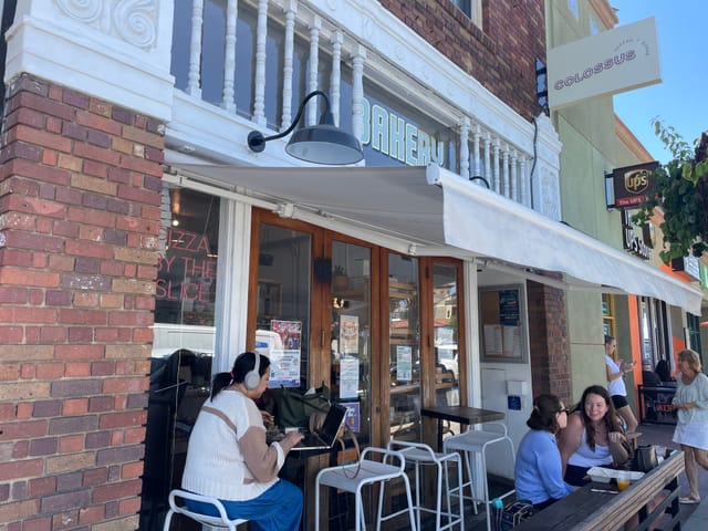 People sitting on white chairs outside a small shop with the word "bakery" printed above the front door.