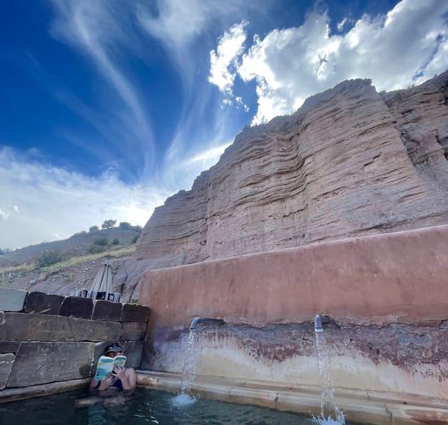 Person in a pool beneath a rocky cliff and bright blue sky.