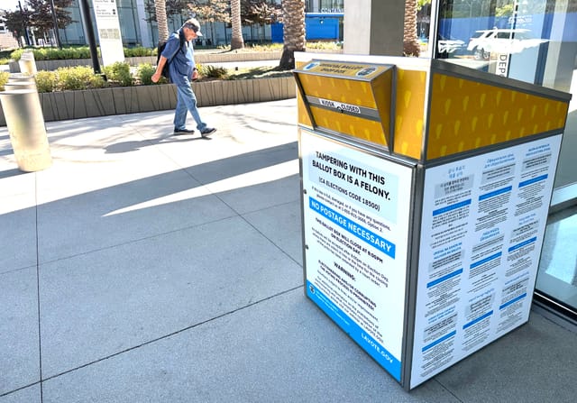 A man walking by a white and yellow ballot box outside an office building.