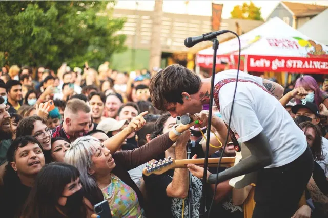 A woman with white hair holds a microphone while a man holding a guitar sings into it.