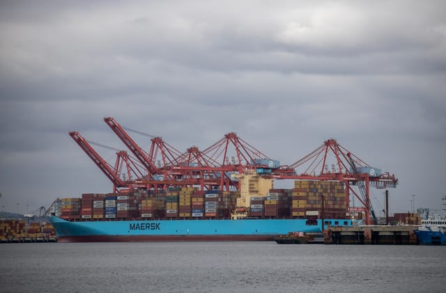 Blue freighter with deck stacked with colorful containers docked near red cranes.