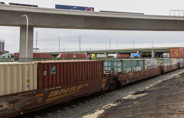 Different colored shipping containers sit on train cars and are pulled behind semi-trucks.