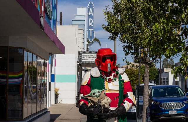 A man wears a red version of a helmet worn by Star Wars villains while holding a small green Star Wars character standing in 