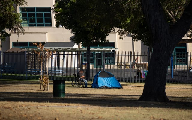 A man sits in a wheelchair next to a blue tent at a park across the street from a school.