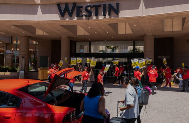 People wearing red shirts and holding signs that say "on strike" stand outside a hotel.