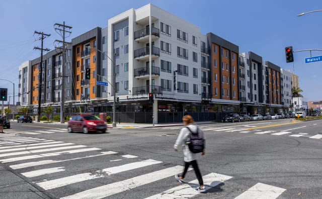 A pedestrian in a crosswalk passes a large white, brown and gray apartment building.