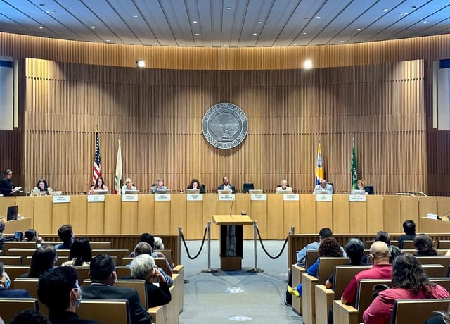 Nine people sit on a beige dais facing a couple dozen people siting in rows of theater seats.