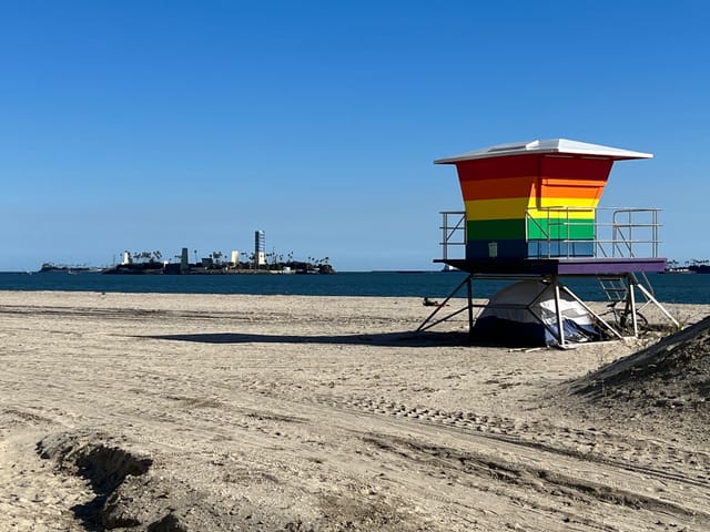 Rainbow colored lifeguard tower on a beach in front of a bright blue ocean and a small island covered with buildings.