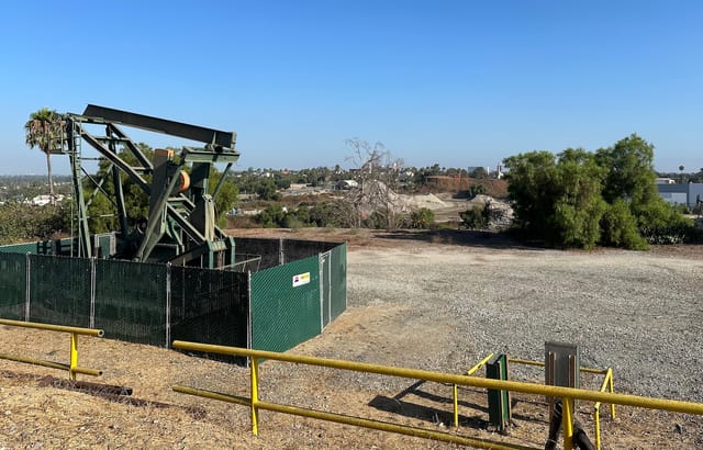 Oil well surrounded by a green fence near an old yellow metal railing in an open field.