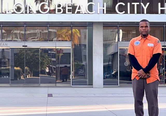 A Black man in an orange shirt with long black sleeves stands in front of a building with the words Long Beach City.