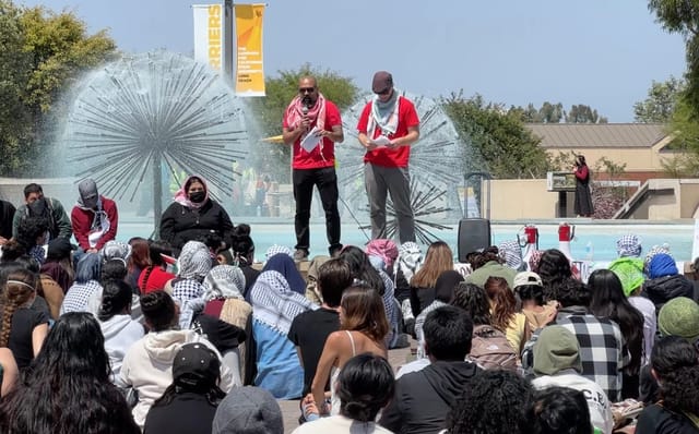 Two men wearing read shirts stand in front of a fountain in front of dozens of people.