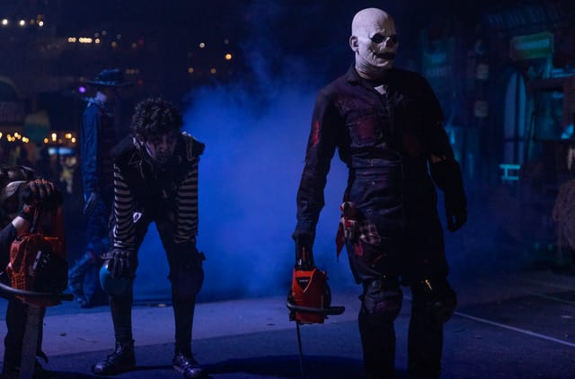 Men in scary costumes and masks stand in a dark, foggy area.