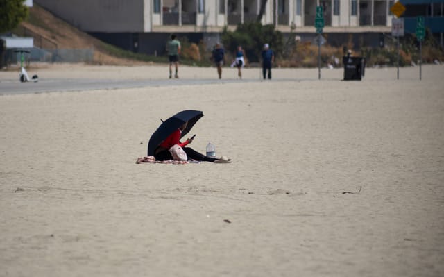 A person wearing a red shirt sits on sand under a large black umbrella.