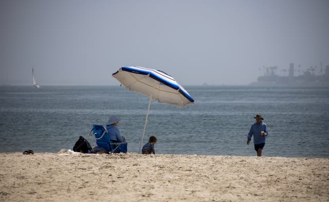 A woman and child sit under a blue and white umbrella on a beach while a man walks toward them, away form the wate