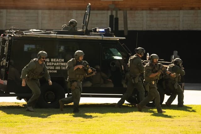 Police officers in body armor run alongside a black armored vehicle.