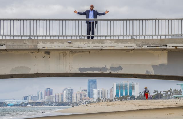 A man stands on a pier with his arms spread with a city skyline visible under the pier in the distance.