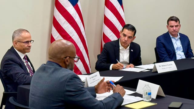 Four men sit at a black table scowling at each other in front of two American flags.