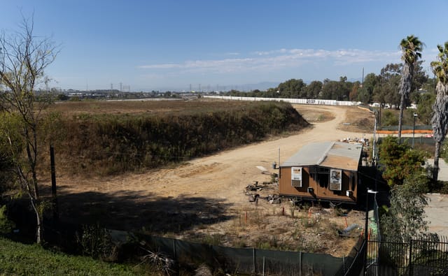 A small brown temporary building sits near a dirt road that borders a large raised earthen moun