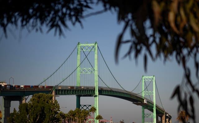 Semi-trucks carry shipping containers over a green suspension bridge.