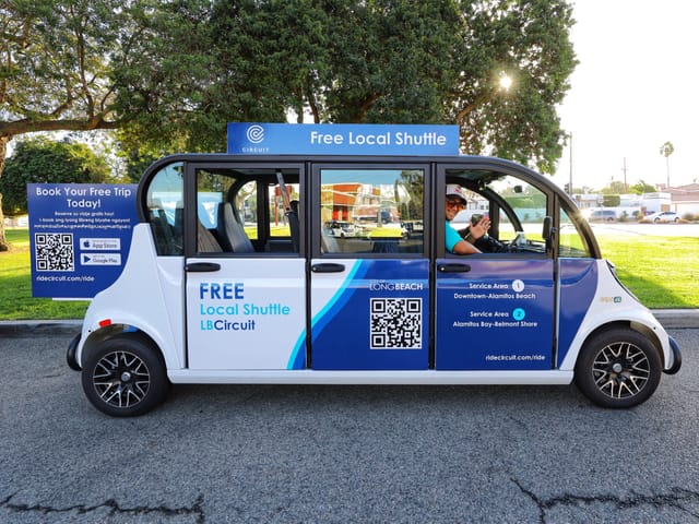 A person smiles in the driver's seat of a tiny white and blue micro-transit vehicle.