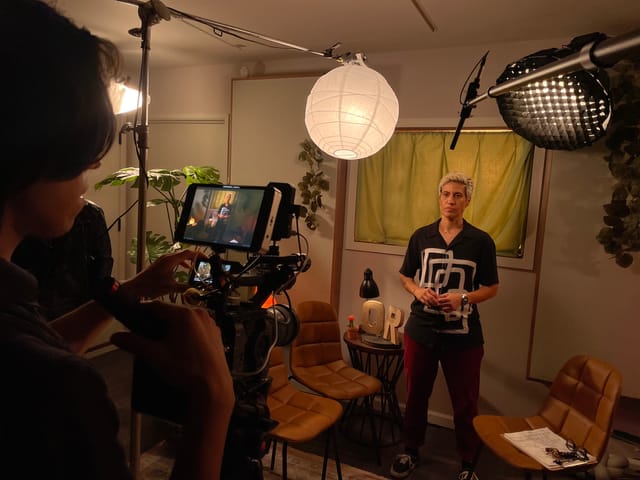 A person stands in a room next to three brown chairs while being filmed by another person.