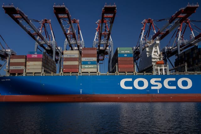 Giant red cranes tower over a ship loaded with multi-colored shipping containers.