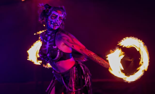 A woman in dramatic face paint twirls a stick with fire on both ends.