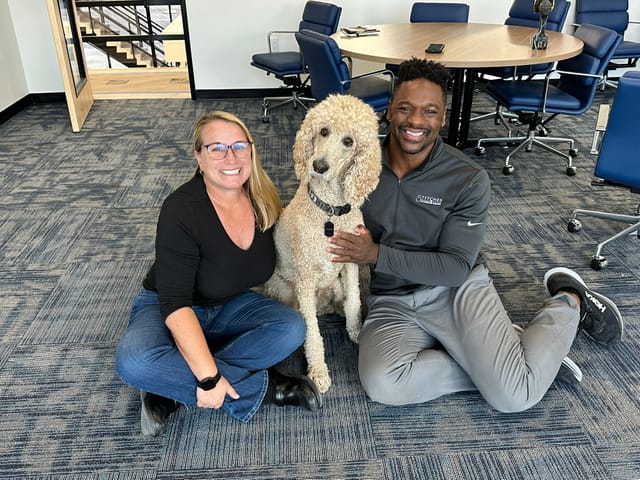 Two people and a large poodle sit on the floor in an otherwise empty office.