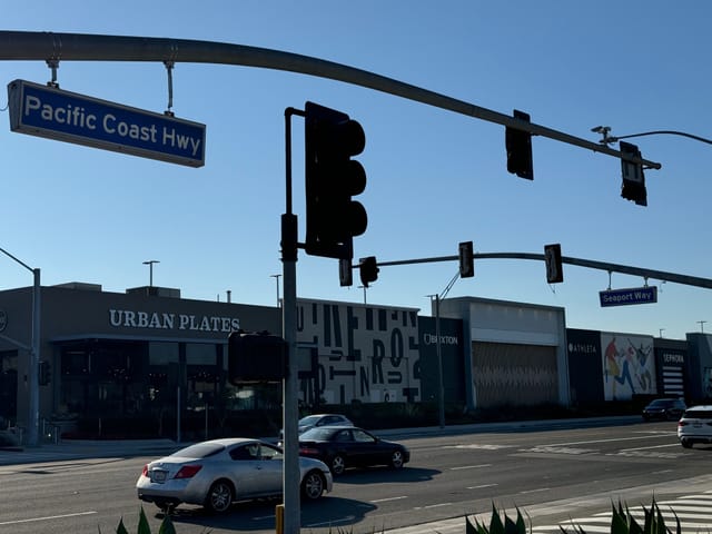 Cars pass by a shopping center in Long Beach, with traffic signals visible over the street.