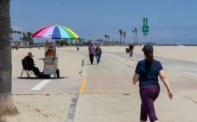 People walk on a concrete beach path by a food vendor cart parked under a rainbow colored umbrella.