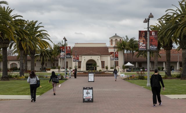 People walk on a brick path in front of palm trees and white buildings with a red tile roof.