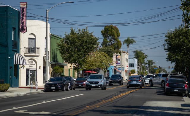 Cars driving past a variety of two-story buildings.