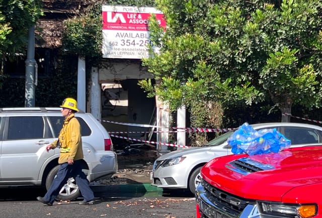 A man wearing a yellow helmet and jacket walks past a building taped off after a fire.