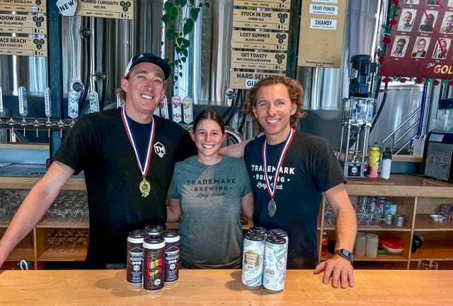 Three smiling people, two of which are wearing gold medals around their necks, standing in front of eight cans of beer.