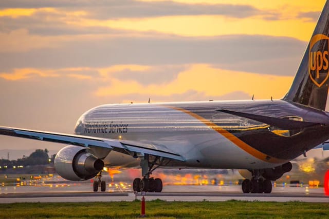 A brown and white plane marked "UPS" taxis on a runway at sunset.