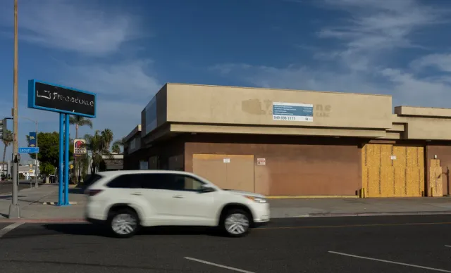 A blurry white car drives past a boarded up building.